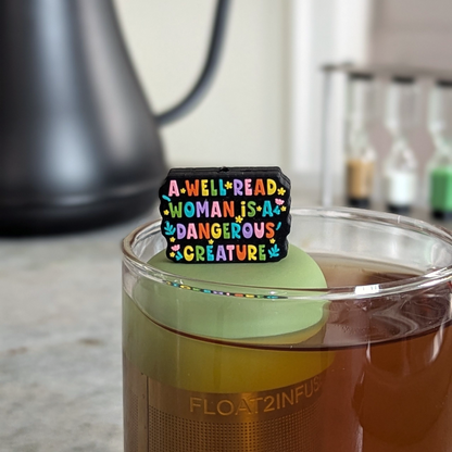 black rectangle with text "a well read woman is a dangerous creature" in rainbow font tea infuser for loose leaf tea  with a fine mesh stainless steel strainer that reads FLOAT2INFUSE on a green base floating in a glass tea mug with a tea kettle in the background