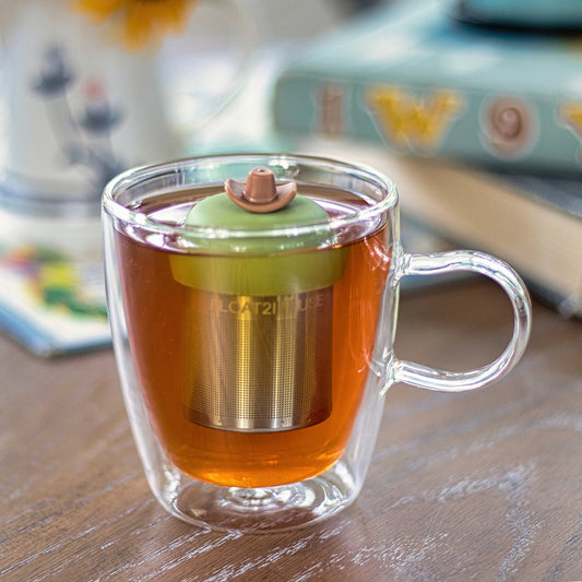 brown cowboy hat tea infuser for loose leaf tea with a fine mesh stainless steel tea strainer that reads FLOAT2INFUSE on a green base  floating in a glass tea mug 
with books in the background