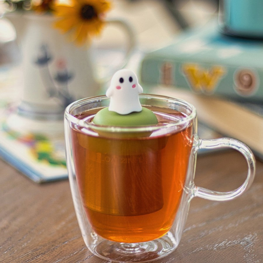 white ghost with cute pink cheeks tea infuser for loose leaf tea with a fine mesh stainless steel tea strainer 
that reads FLOAT2INFUSE on a green base floating in a glass tea mug 
with books in the background