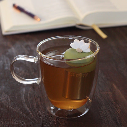 white lotus flower tea infuser for loose leaf tea  with a fine mesh stainless steel tea infuser that reads FLOAT2INFUSE on a green base floating in a glass tea mug with notebook and pen in the background