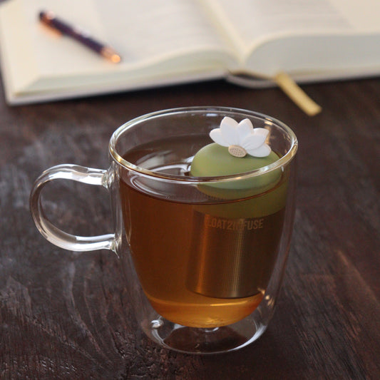 white lotus flower tea infuser for loose leaf tea  with a fine mesh stainless steel tea infuser that reads FLOAT2INFUSE on a green base floating in a glass tea mug with notebook and pen in the background