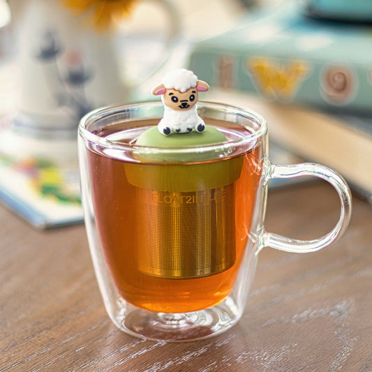 happy white sheep animal  tea infuser for loose leaf tea with a fine mesh stainless steel tea strainer that reads FLOAT2INFUSE on a green base  floating in a glass tea mug with books in the background