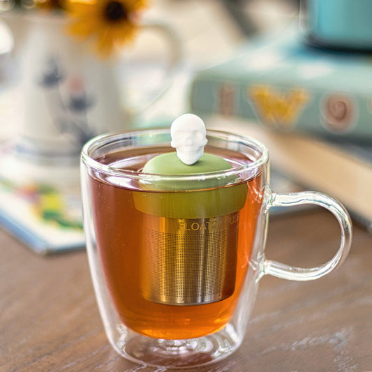 white skull tea infuser for loose leaf tea with a fine mesh stainless steel tea strainer that reads FLOAT2INFUSE on a green base  floating in a glass tea mug 
with books in the background
