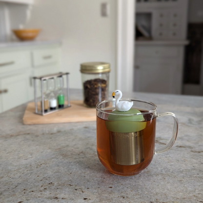 white swan tea infuser  for loose leaf tea with fine mesh stainless steel tea strainer that reads FLOAT2INFUSE on a green base floating in a glass tea mug with loose leaf tea in the background
