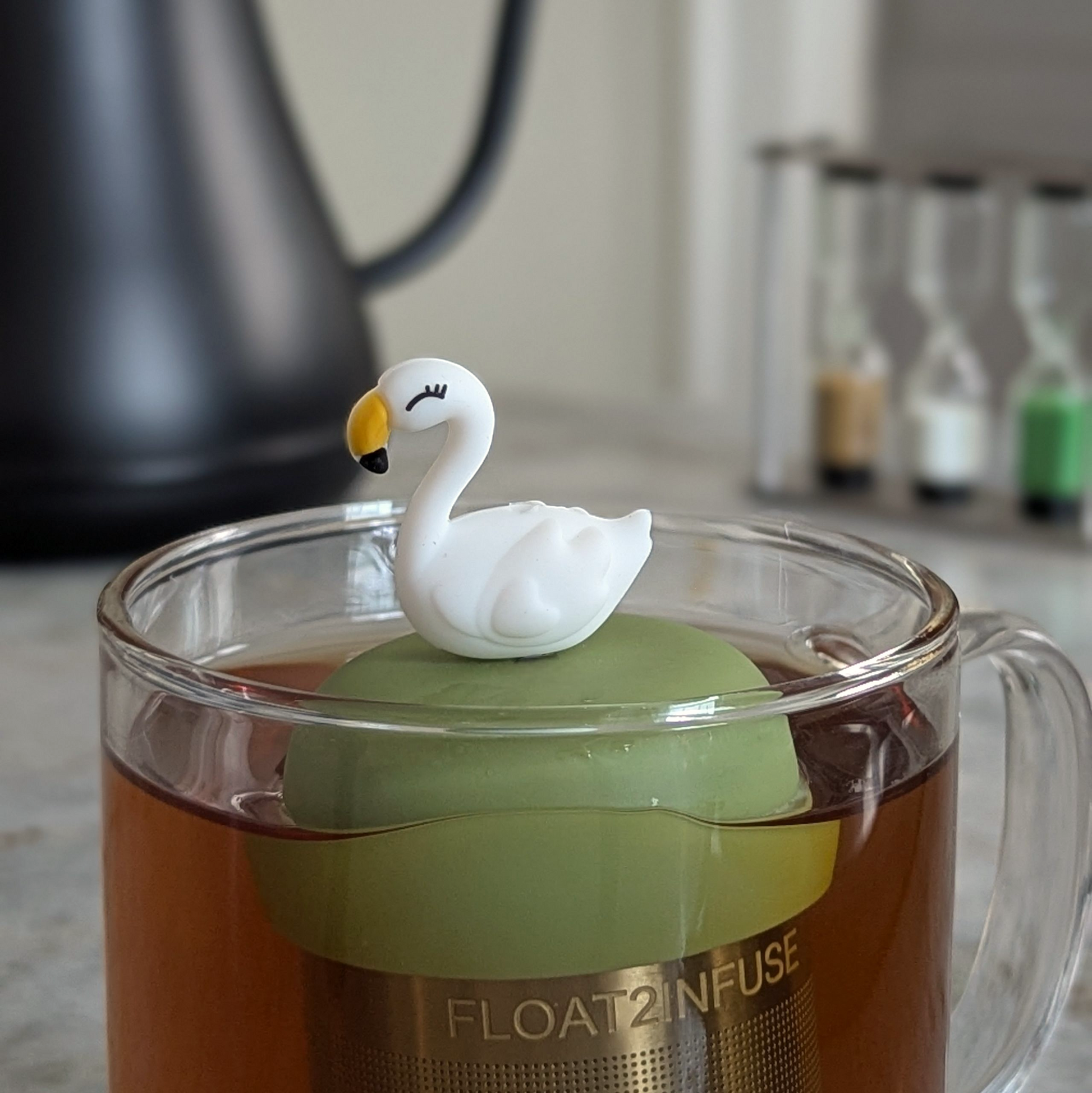 white swan tea infuser  for loose leaf tea with fine mesh stainless steel tea strainer that reads FLOAT2INFUSE on a green base floating in a glass tea mug with a tea kettle in the background