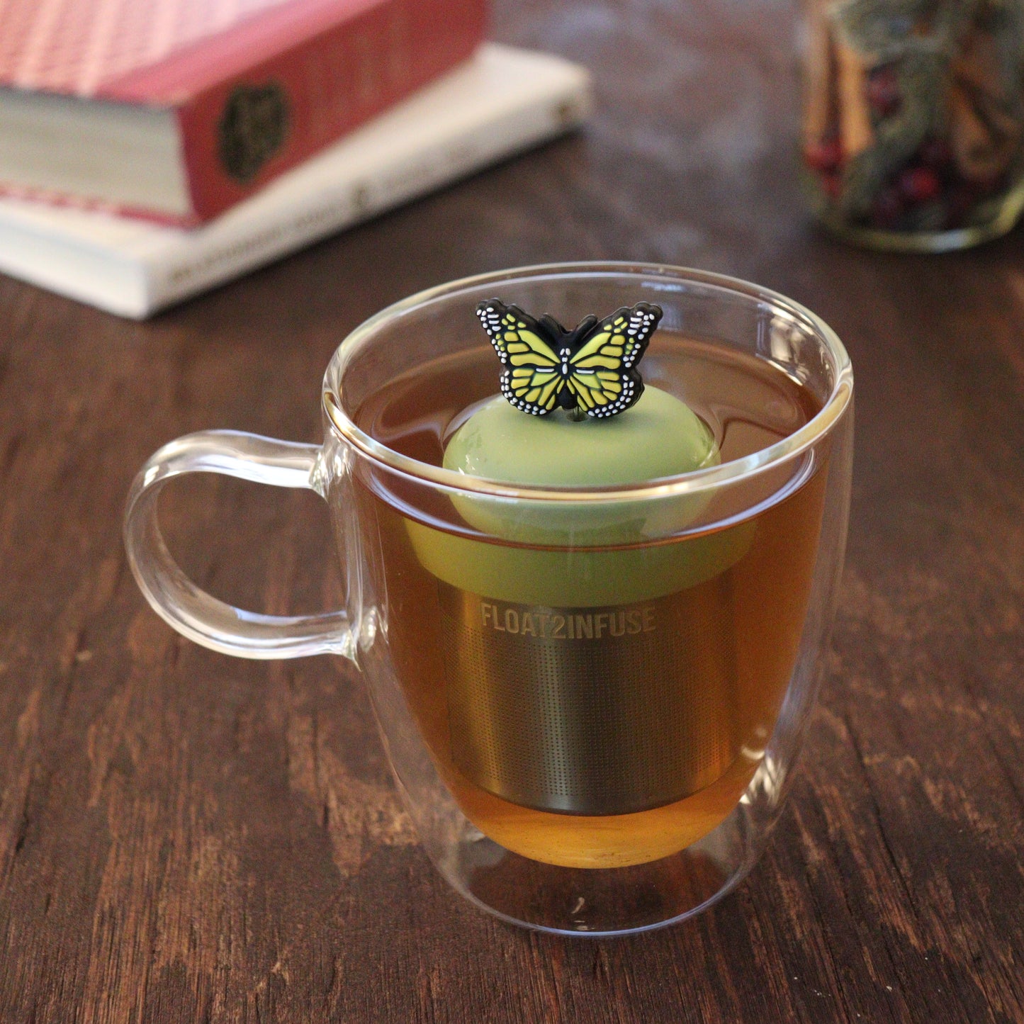 yellow butterfly tea infuser for loose leaf tea  with a fine mesh stainless steel tea infuser that reads FLOAT2INFUSE on a green base floating in a glass tea mug with books in the background
