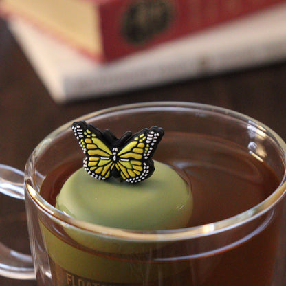 Yellow butterfly tea infuser for loose leaf tea with a fine mesh stainless steel tea strainer that reads FLOAT2INFUSE on a green base floating in a glass tea mug with books in the background 