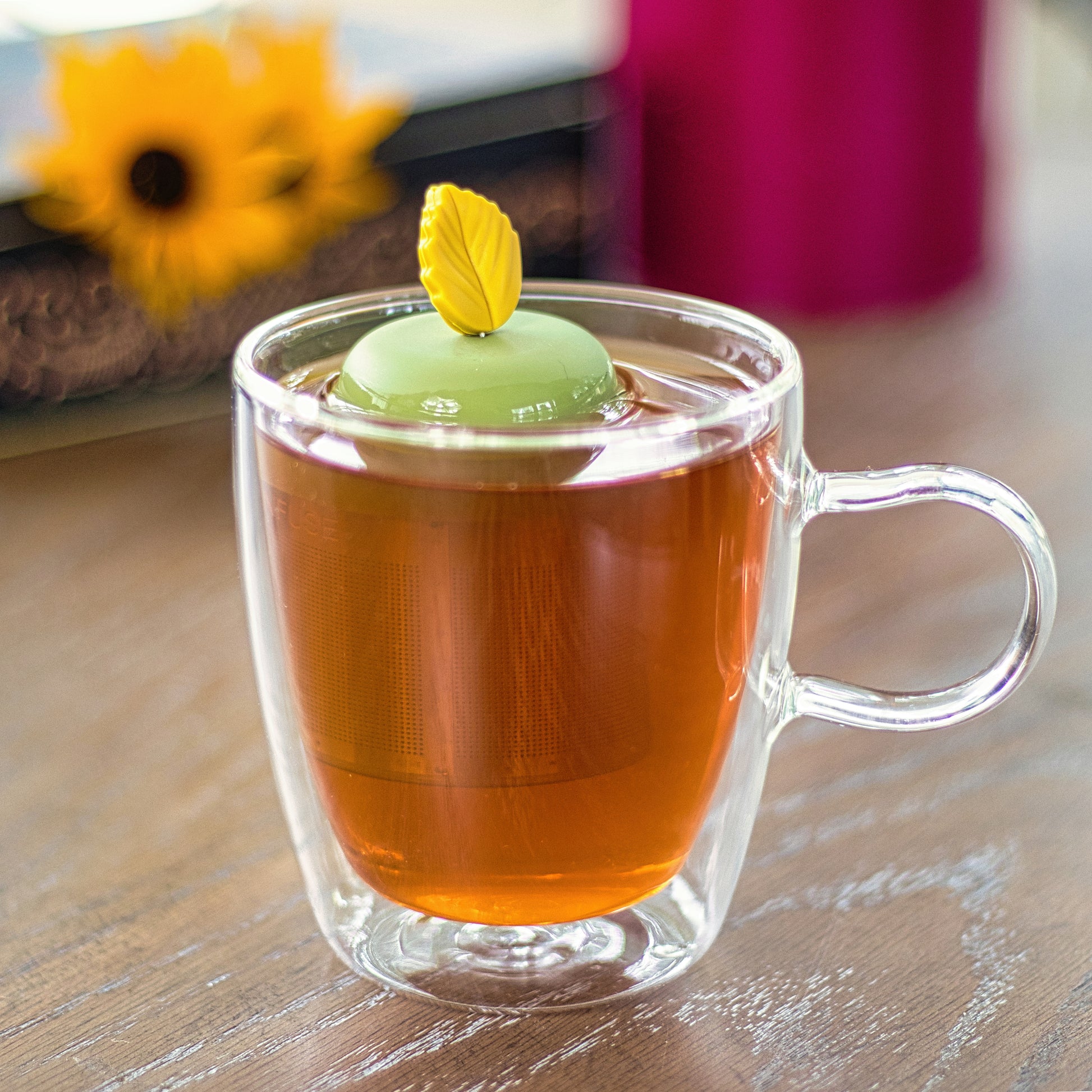 yellow fall leaf tea infuser for loose leaf tea with a fine mesh stainless steel tea strainer that reads FLOAT2INFUSE on a green base  floating in a glass tea mug with flowers in the background