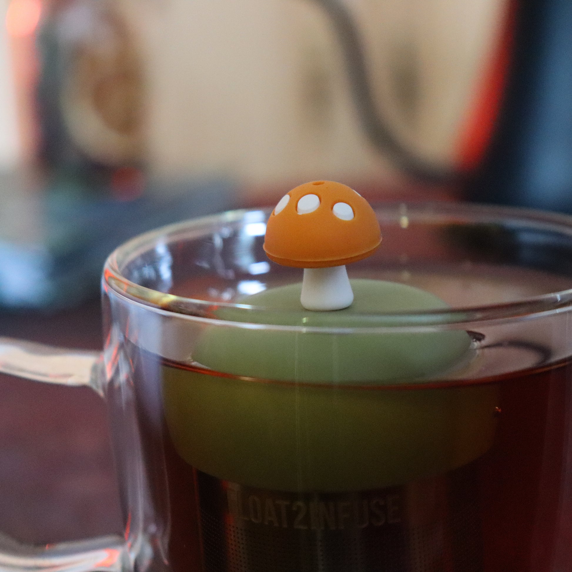 yellow mushroom tea infuser for loose leaf tea  with a fine mesh stainless steel tea strainer that reads FLOAT2INFUSE on a green base floating in a glass tea mug with tea kettle in the background