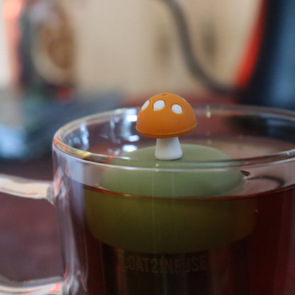 yellow mushroom tea infuser for loose leaf tea  with a fine mesh stainless steel tea strainer that reads FLOAT2INFUSE on a green base floating in a glass tea mug with tea kettle in the background
