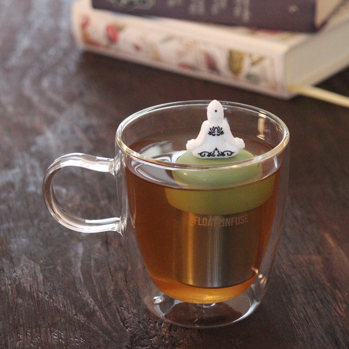 white yogi tea infuser for loose leaf tea  with a fine mesh stainless steel tea infuser that reads FLOAT2INFUSE on a green base floating in a glass tea mug with books in the background