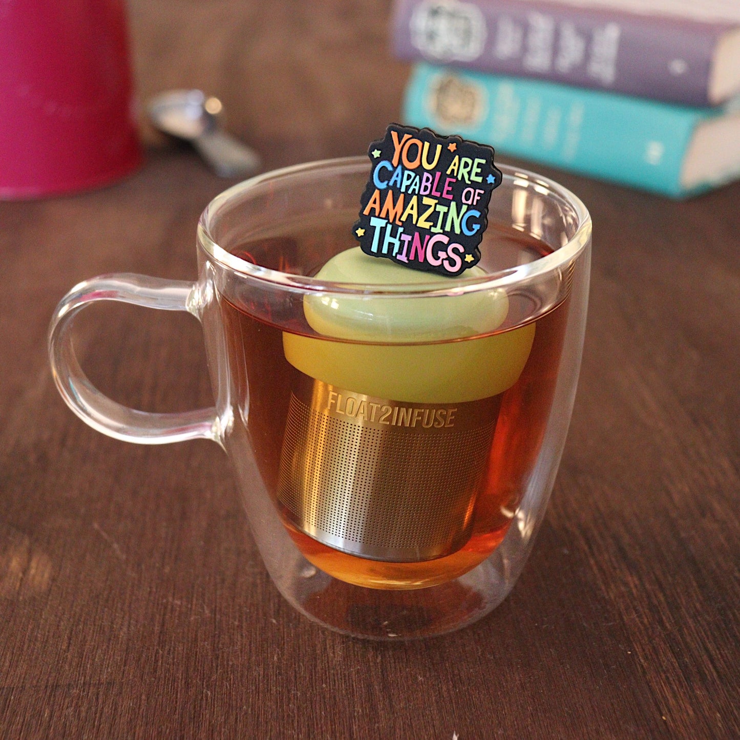 Text that reads "You are capable of amazing things" in bright rainbow font tea infuser for loose leaf tea with a fine mesh stainless steel tea strainer that reads FLOAT2INFUSE on a green base with a tea canister and books in the background