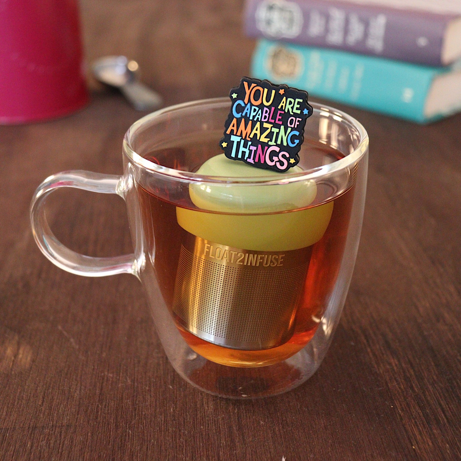 Text that reads "You are capable of amazing things" in bright rainbow font tea infuser for loose leaf tea with a fine mesh stainless steel tea strainer that reads FLOAT2INFUSE on a green base with a tea canister and books in the background
