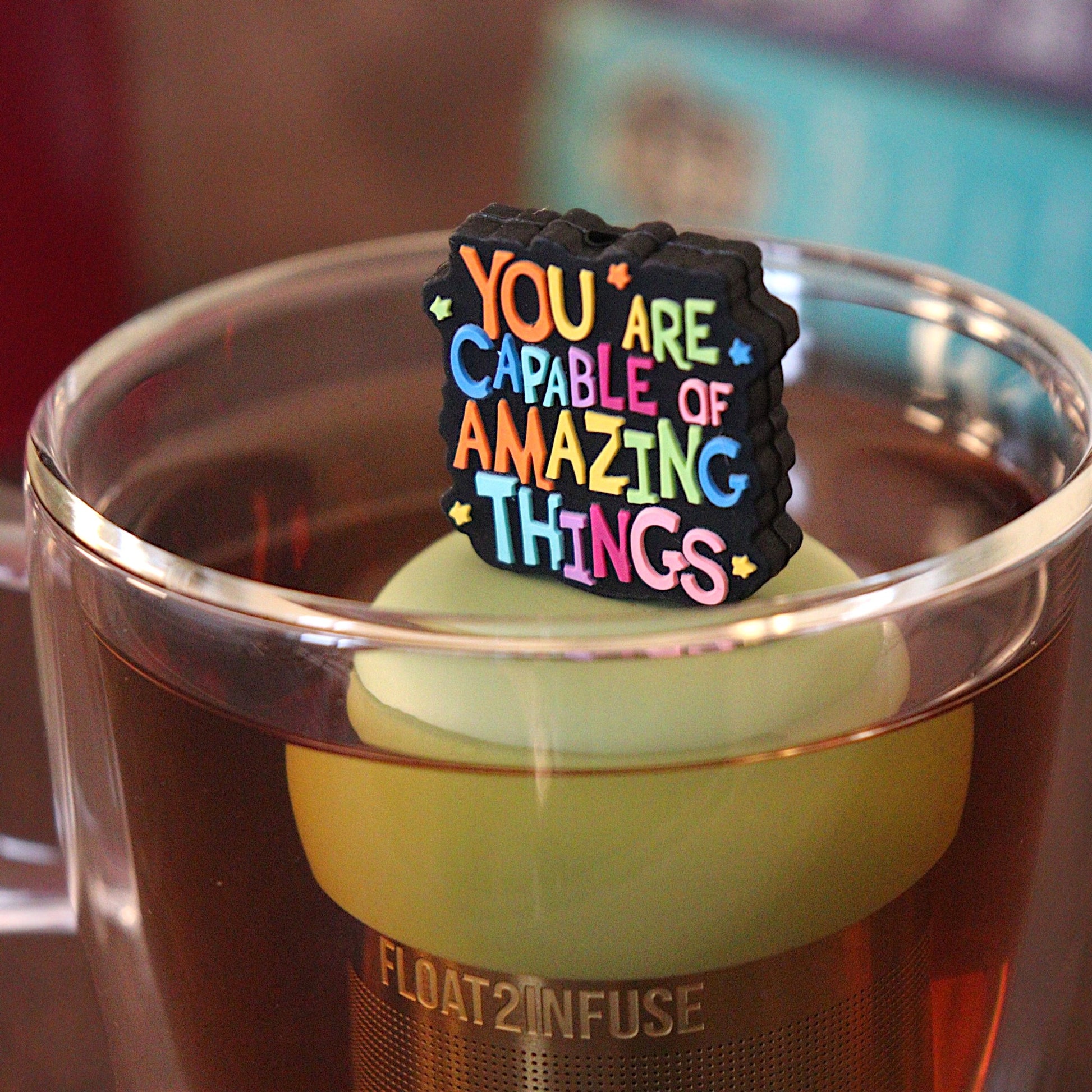 Text that reads "You are capable of amazing things" in bright rainbow font tea infuser for loose leaf tea with a fine mesh stainless steel tea strainer that reads FLOAT2INFUSE on a green base with a tea canister and books in the background