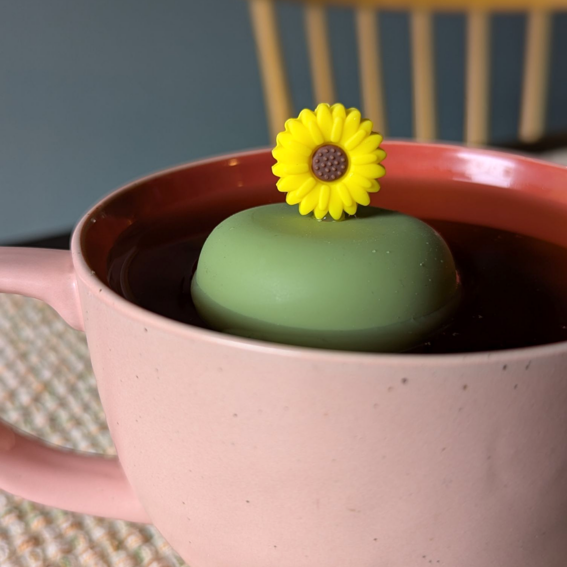 yellow sunflower tea infuser for loose leaf tea with a fine mesh stainless steel tea strainer that reads FLOAT2INFUSE on a green base  floating in a pink tea mug with a chair in the background
