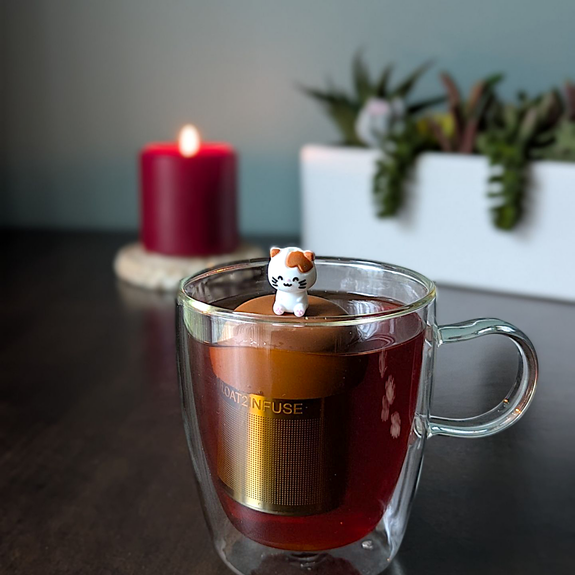 Clear mug with a tea infuser shaped like a cat, candle, and plant in the background